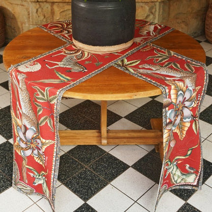 Two red Ardmore wildlife table runners crossed over a round wooden table with a large black decorative vessel at the center.