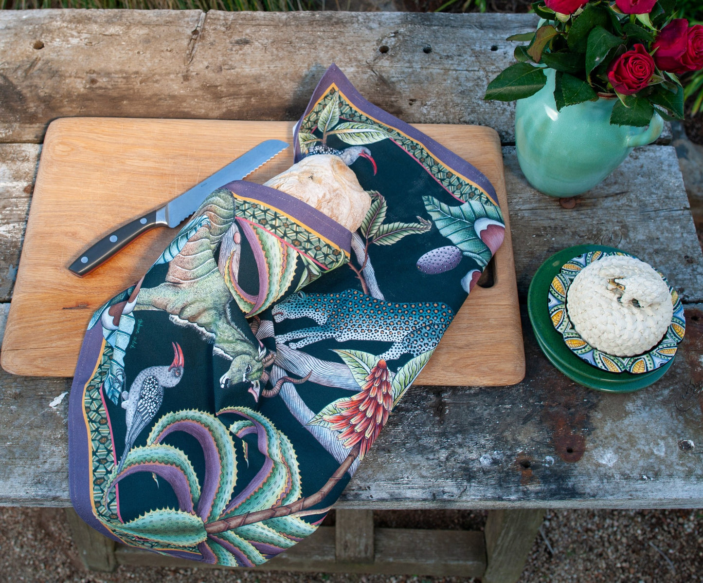 Ardmore wildlife-print tea towel wrapped around a loaf of bread on a rustic outdoor table with a cutting board, knife, flowers, and small dishes.