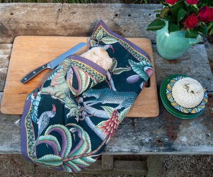 Ardmore wildlife-print tea towel wrapped around a loaf of bread on a rustic outdoor table with a cutting board, knife, flowers, and small dishes.