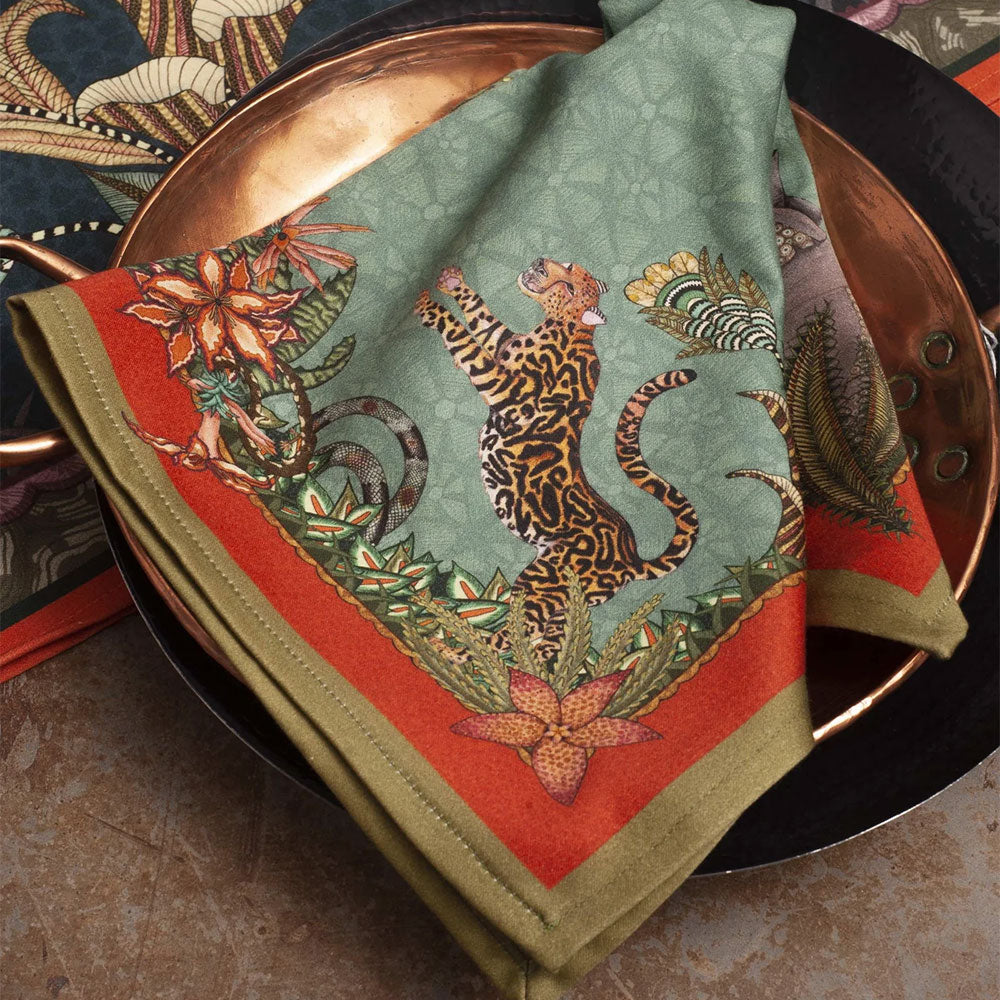Sabie Forest Napkin styled over layered copper and black tableware, featuring a tiger surrounded by tropical foliage on an aqua background with coral and olive green borders.