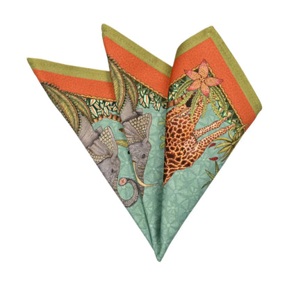 Sabie Forest Napkin folded into a decorative fan shape, showing illustrated elephants, giraffe, tropical foliage, and coral and olive green border accents on an aqua background.