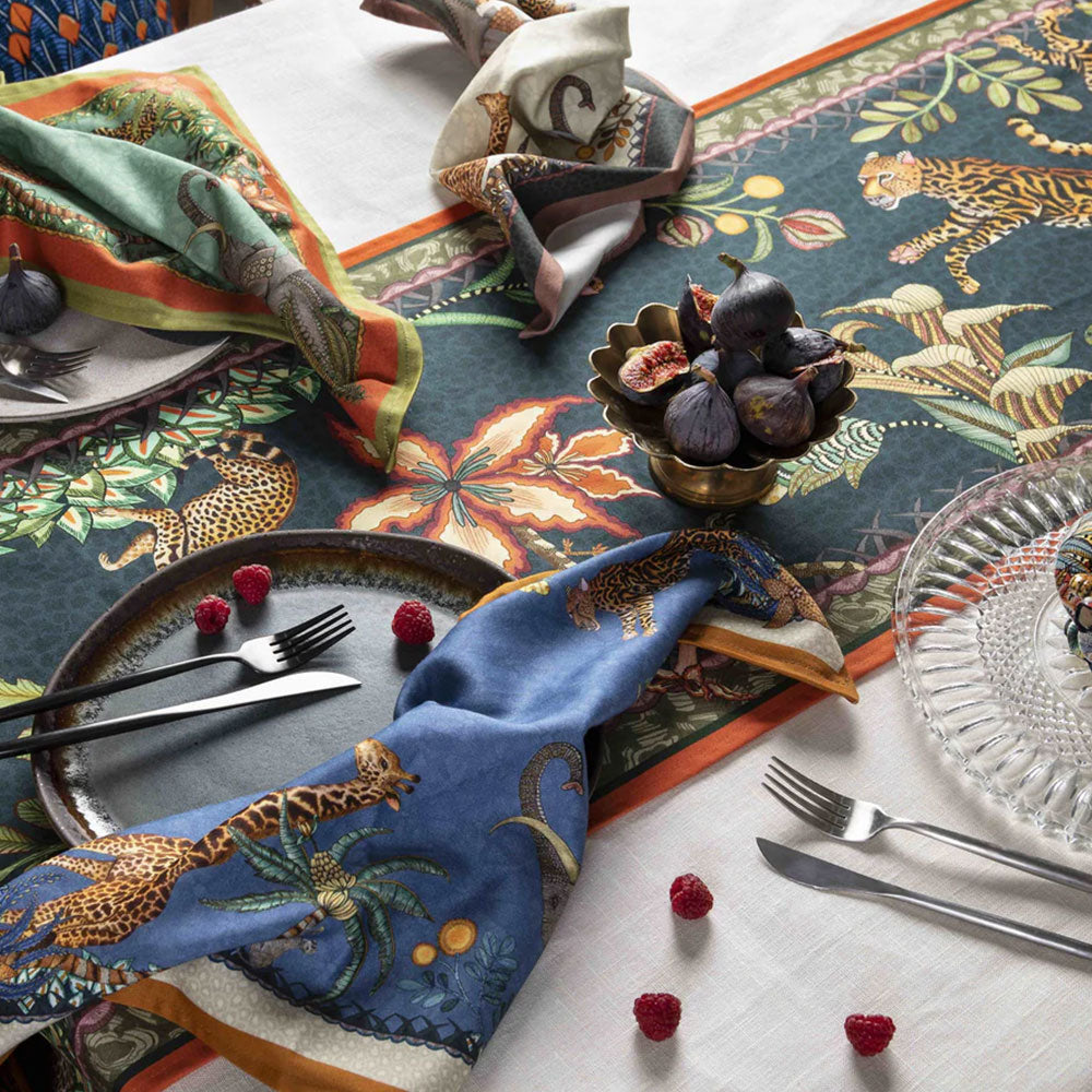 A styled dining table setting featuring the Sabie Forest Table Runner Large in dark teal laid over a white tablecloth, decorated with a brass bowl of figs, scattered raspberries, tableware, and patterned cloth napkins.