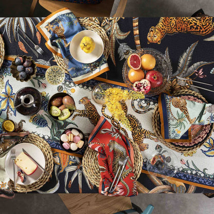 An overhead view of a styled dining table layered with the light-colored Sabie Forest Table Runner Small over a dark safari tablecloth, decorated with woven placemats, fresh fruits, macarons, and patterned cloth napkins.