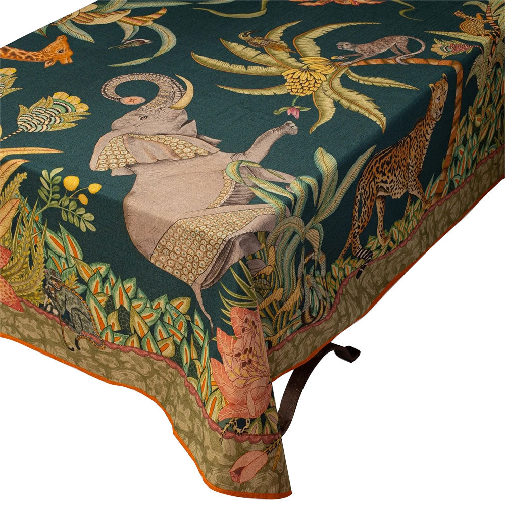 Close-up corner view of the Sabie Forest Tablecloth Small in dark green draped over a table, highlighting an illustrated elephant with raised trunk alongside tropical palm trees, a leopard, and an orange-trimmed border.