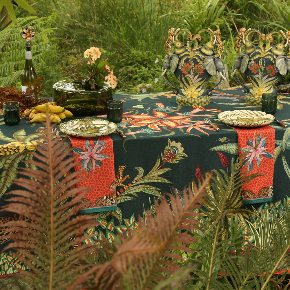 An outdoor dining table set with the dark green Sabie Forest Tablecloth Small, featuring patterned red and teal napkins, ceramic plates, green drinking glasses, decorative elephant-handled urns, wine, and tropical greenery in the foreground.