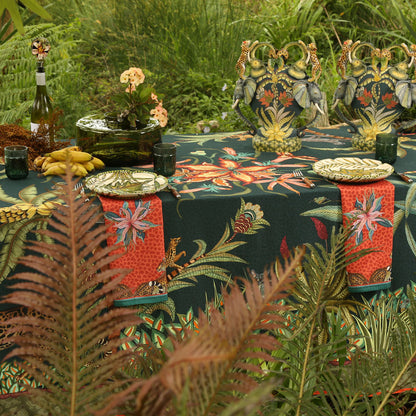 An outdoor dining table set with the dark green Sabie Forest Tablecloth Small, featuring patterned red and teal napkins, ceramic plates, green drinking glasses, decorative elephant-handled urns, wine, and tropical greenery in the foreground.