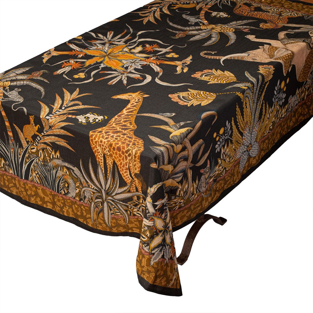 A rectangular table covered with the black Sabie Forest Tablecloth, featuring detailed illustrations of a giraffe, foliage, and safari animals draped neatly over the edges, surrounded by a decorative brown border.
