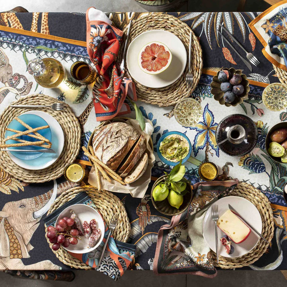 An overhead view of a fully set dining table using the Sabie Forest Tablecloth, featuring woven placemats, white plates with fresh fruit and cheese, sliced bread, dipping dishes, glassware, and cloth napkins.