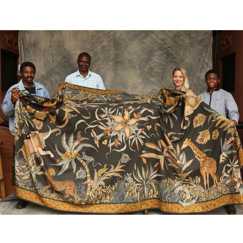 Four people standing indoors holding up the large Sabie Forest Tablecloth in black to show its full size, scale, and detailed safari pattern featuring giraffes, elephants, and tropical plants.