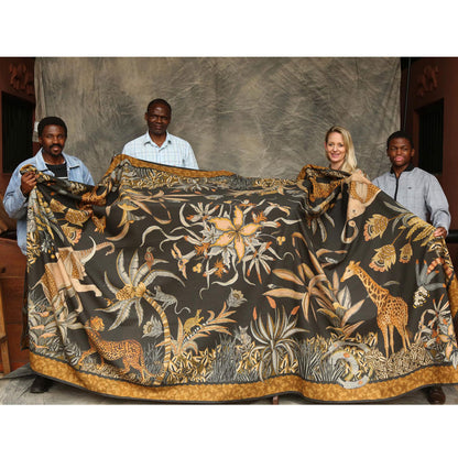 Four people standing indoors holding up the large Sabie Forest Tablecloth in black to show its full size, scale, and detailed safari pattern featuring giraffes, elephants, and tropical plants.