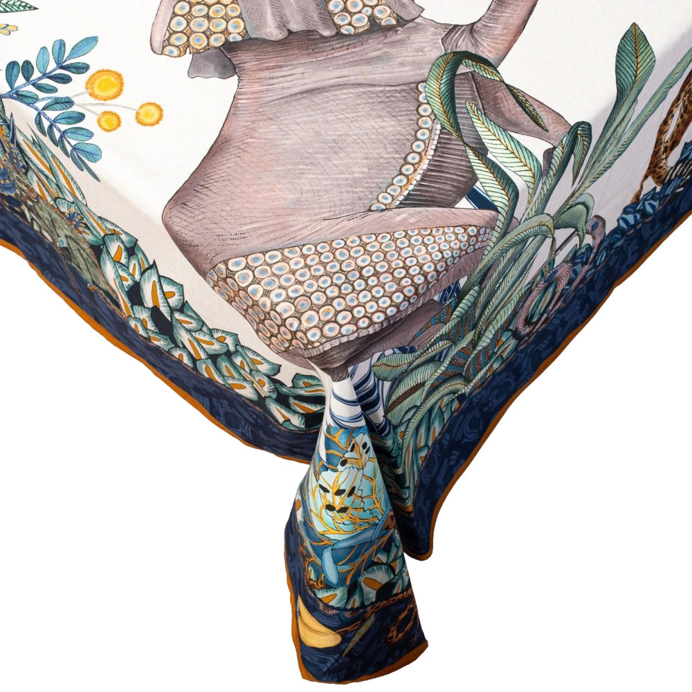 Close-up of a corner of the Sabie Forest tablecloth hanging off a table edge, highlighting the detailed illustration of an elephant with decorative patterned skin, surrounded by lush green foliage, a blue border, and a gold trimmed edge.