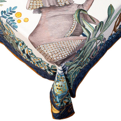Close-up of a corner of the Sabie Forest tablecloth hanging off a table edge, highlighting the detailed illustration of an elephant with decorative patterned skin, surrounded by lush green foliage, a blue border, and a gold trimmed edge.