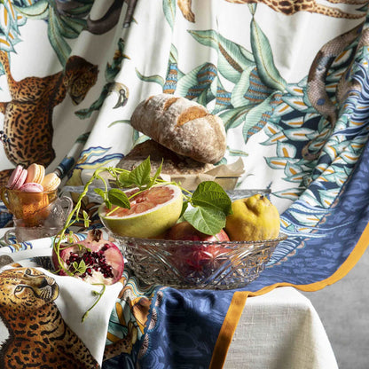Still life arrangement on a table style with the Sabie Forest tablecloth, featuring a glass bowl filled with fresh fruit including a halved grapefruit, pomegranate, and quince, flanked by artisanal bread and a small dish of pastel macarons.