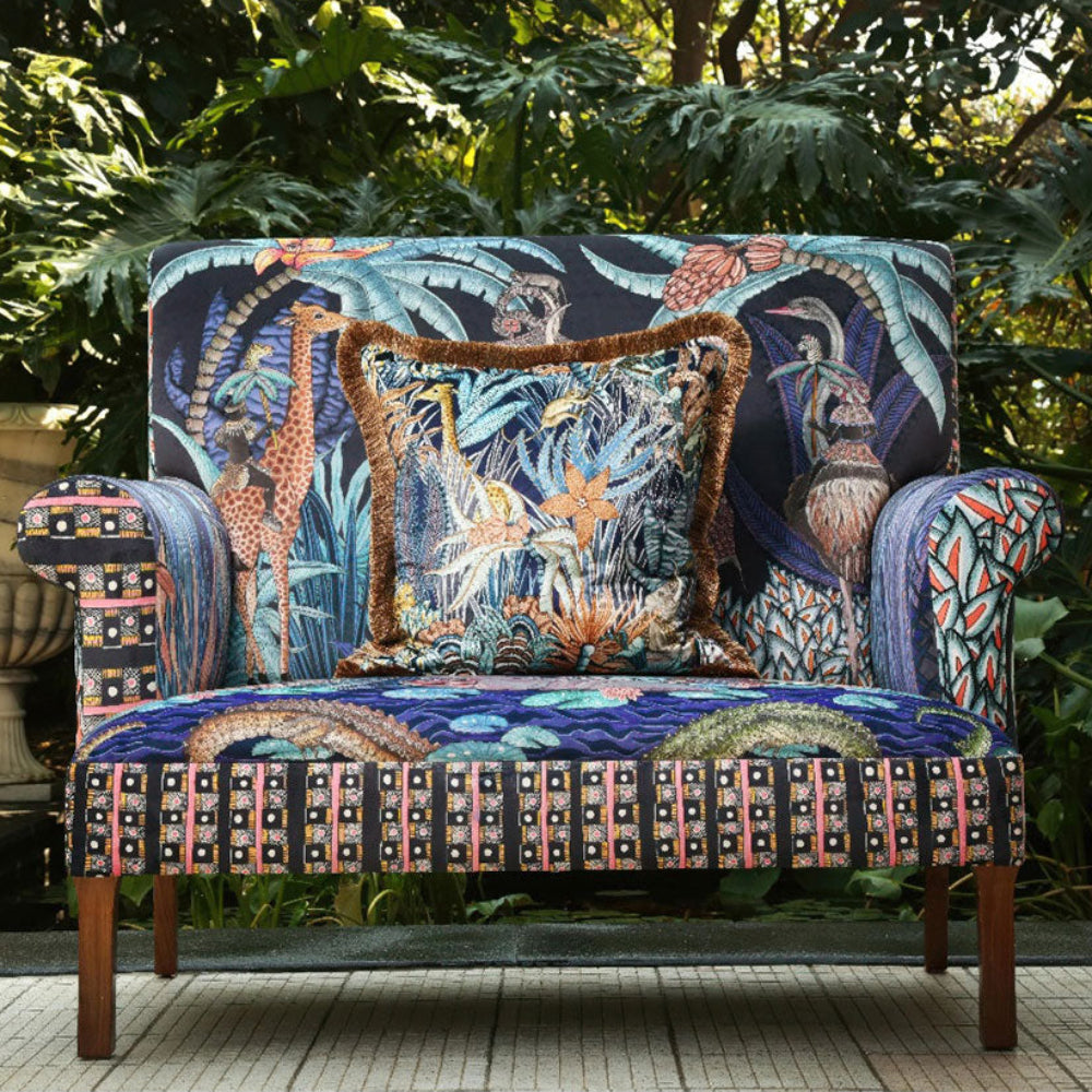 The Sabie Sofa styled outdoors in a lush garden setting with a matching fringe-trimmed decorative throw pillow placed on the seat.
