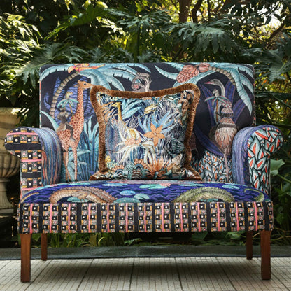 The Sabie Sofa styled outdoors in a lush garden setting with a matching fringe-trimmed decorative throw pillow placed on the seat.