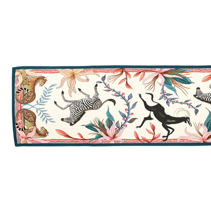 Full horizontal view of the Sabie Forest Table Runner Small laid flat, featuring a cream base with illustrations of leopards, a prancing zebra, a black antelope, and vibrant floral vines enclosed by a teal border.