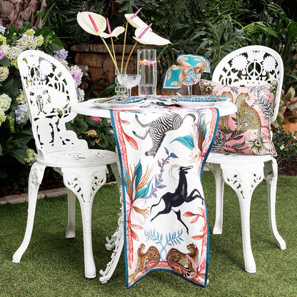 An outdoor garden setting featuring the Sabie Forest Table Runner Small draped across a white wrought-iron patio table with matching chairs, set with glassware, pink anthurium flowers, and a leopard-print cushion.