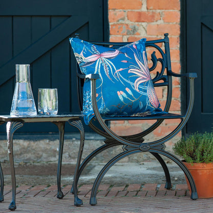 The ocean blue Thanda Nest Pillow styled outdoors on a black wrought-iron armchair beside a matching side table with glassware.