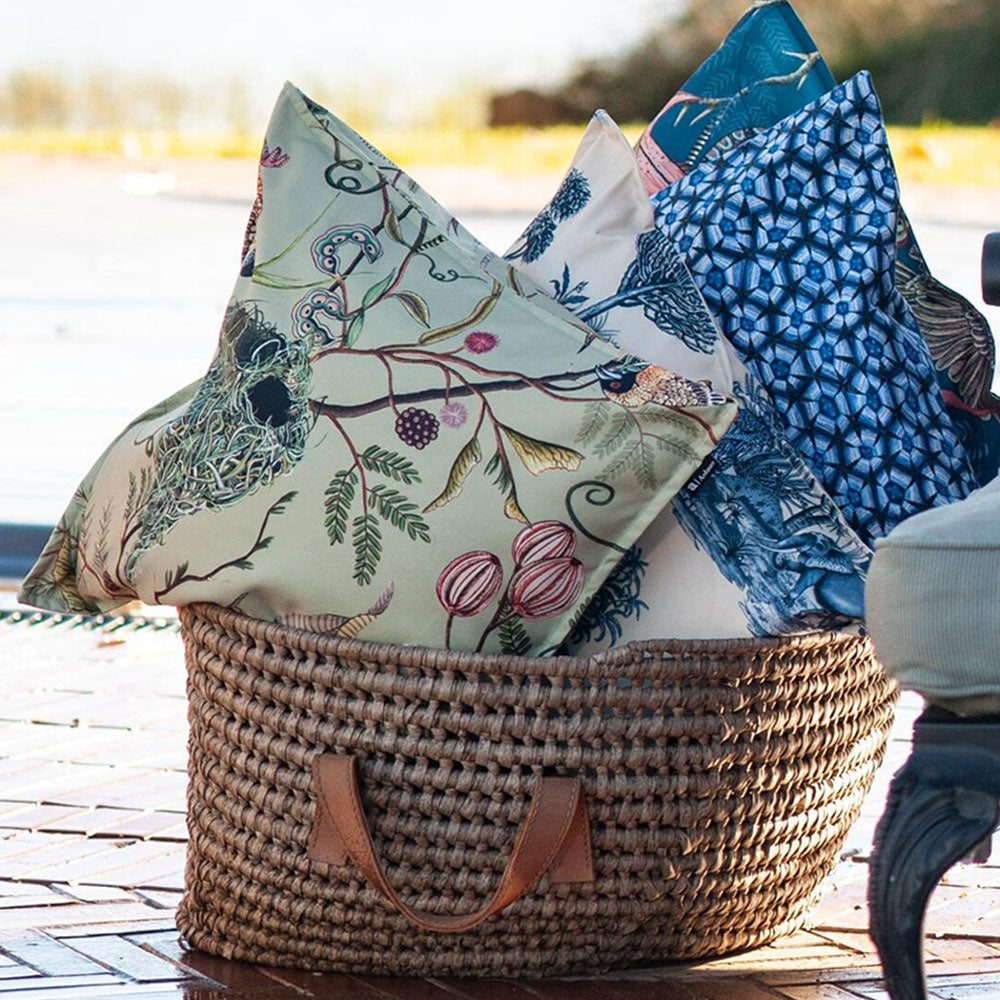 The sage green Thanda Nest Pillow styled inside a woven storage basket alongside blue and white decorative throw pillows outdoors.