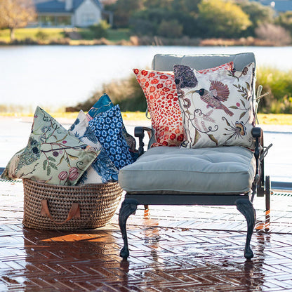 The cream Thanda Nest Pillow styled outdoors on a grey patio lounge chair alongside a basket filled with decorative throw pillows by a lake.