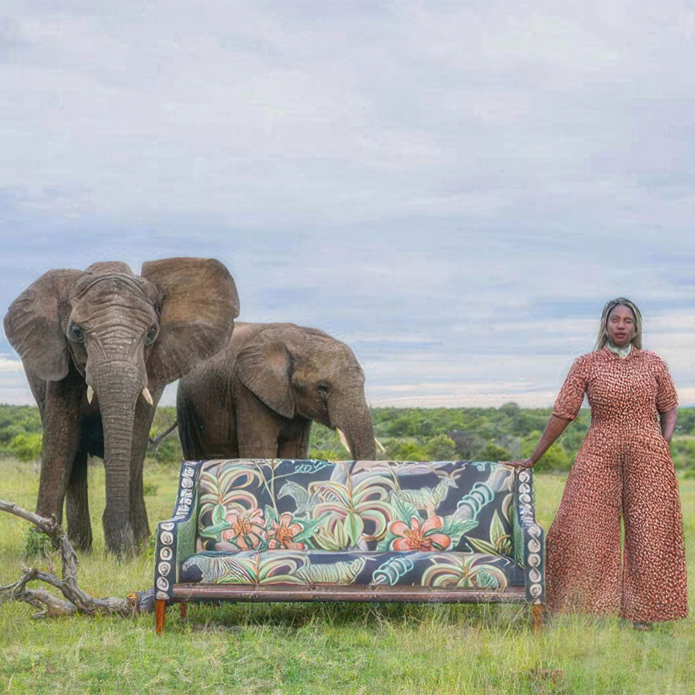 An outdoor photo shoot featuring the Thanda Sofa in a grassy African landscape, posed beside a woman in a patterned jumpsuit and two wild elephants under an open sky.