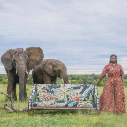 An outdoor photo shoot featuring the Thanda Sofa in a grassy African landscape, posed beside a woman in a patterned jumpsuit and two wild elephants under an open sky.