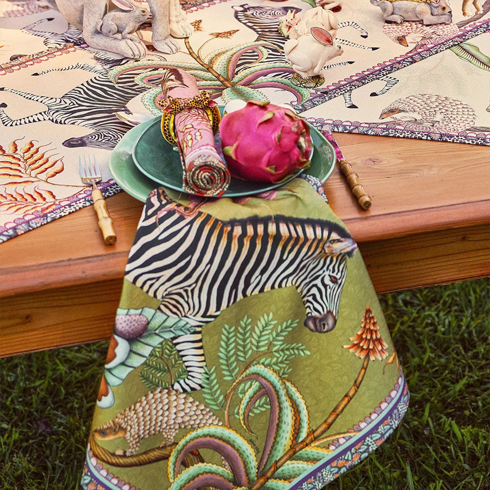 Thanda Stripe Napkin styled on an outdoor dining table, featuring a zebra and botanical print in olive green, cream, pink, and coral beside layered tableware and colorful decorative accents.