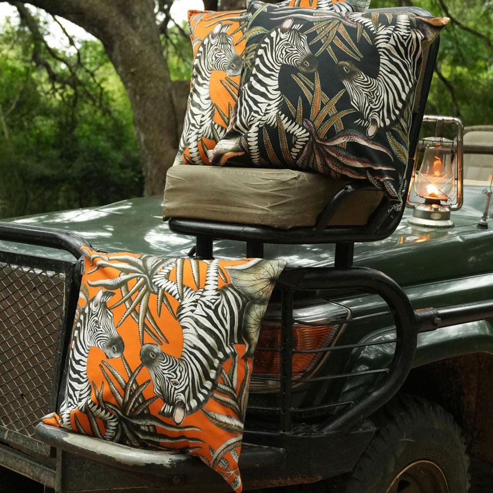 Thanda Stripe Pillows styled on a safari vehicle, showing both deep navy and orange zebra botanical designs in an outdoor setting with a lantern and surrounding greenery.