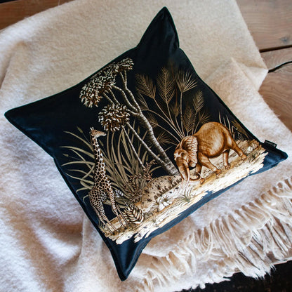 The black Thanda Toile Pillow resting angled on a cozy, cream-colored fringe throw blanket over a wooden surface.