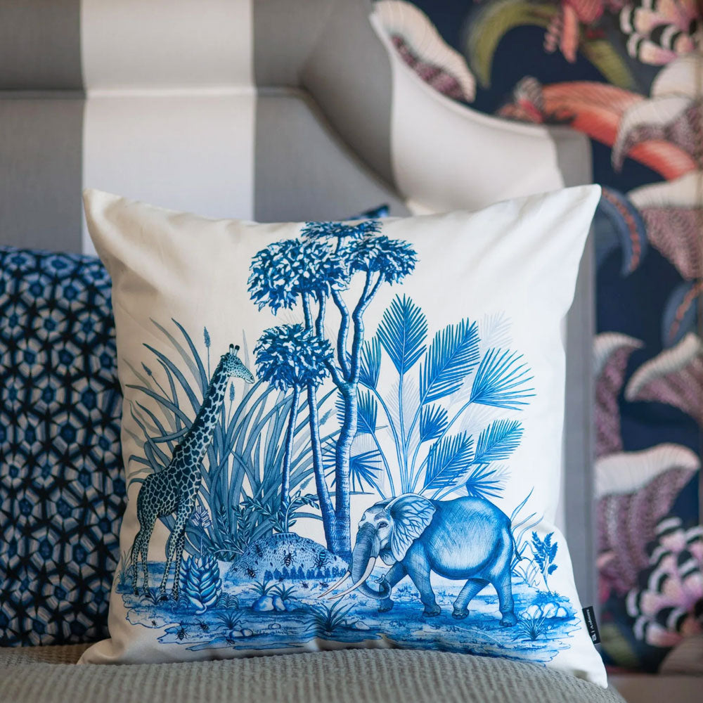 The cream Thanda Nest Pillow styled outdoors on a grey patio lounge chair alongside a basket filled with decorative throw pillows by a lake.