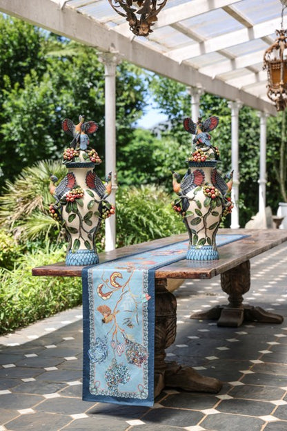 Pale blue Ardmore bird-and-botanical table runner displayed on an outdoor wooden table between two decorative floral vases.